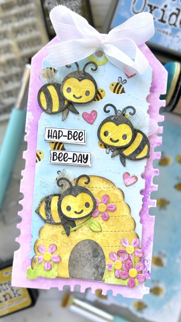 Tag with a watercolored stenciled background with die cut bees and a hive in the foreground